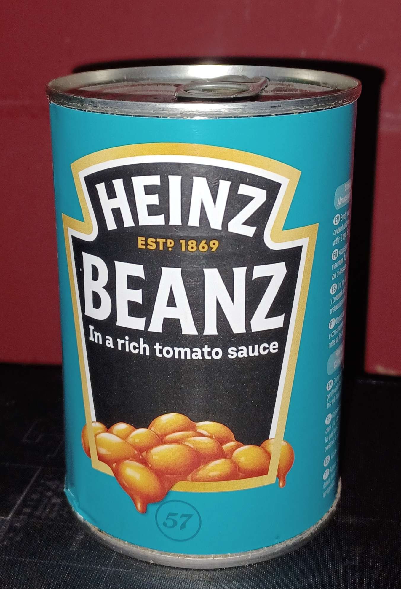 Fotografía real de una lata turquesa Heinz Beanz con desgaste, sobre una superficie oscura y ante un fondo rojo