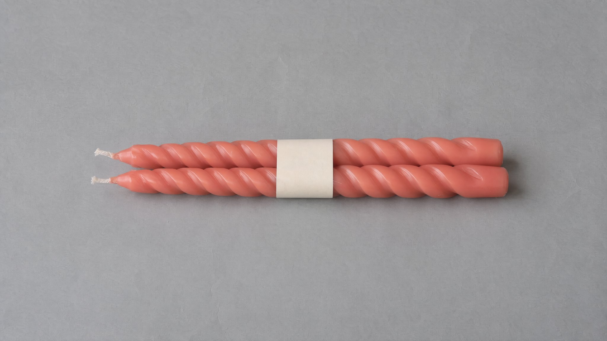 Dos velas coral en espiral con una banda de papel crema sobre papel gris, generadas con IA