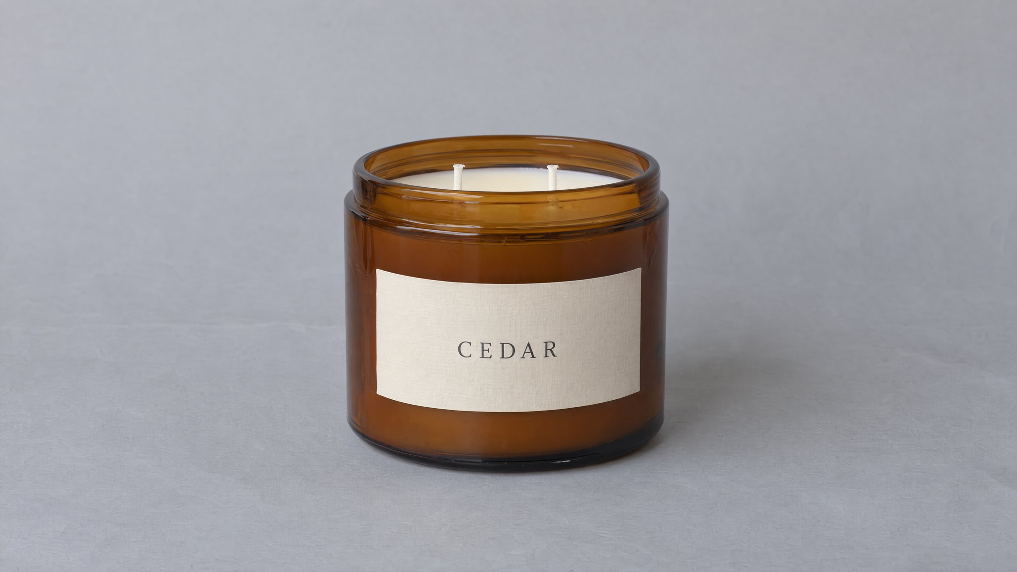Vela CEDAR apagada en recipiente de vidrio ámbar con cera crema y dos mechas sobre papel gris, generada con IA
