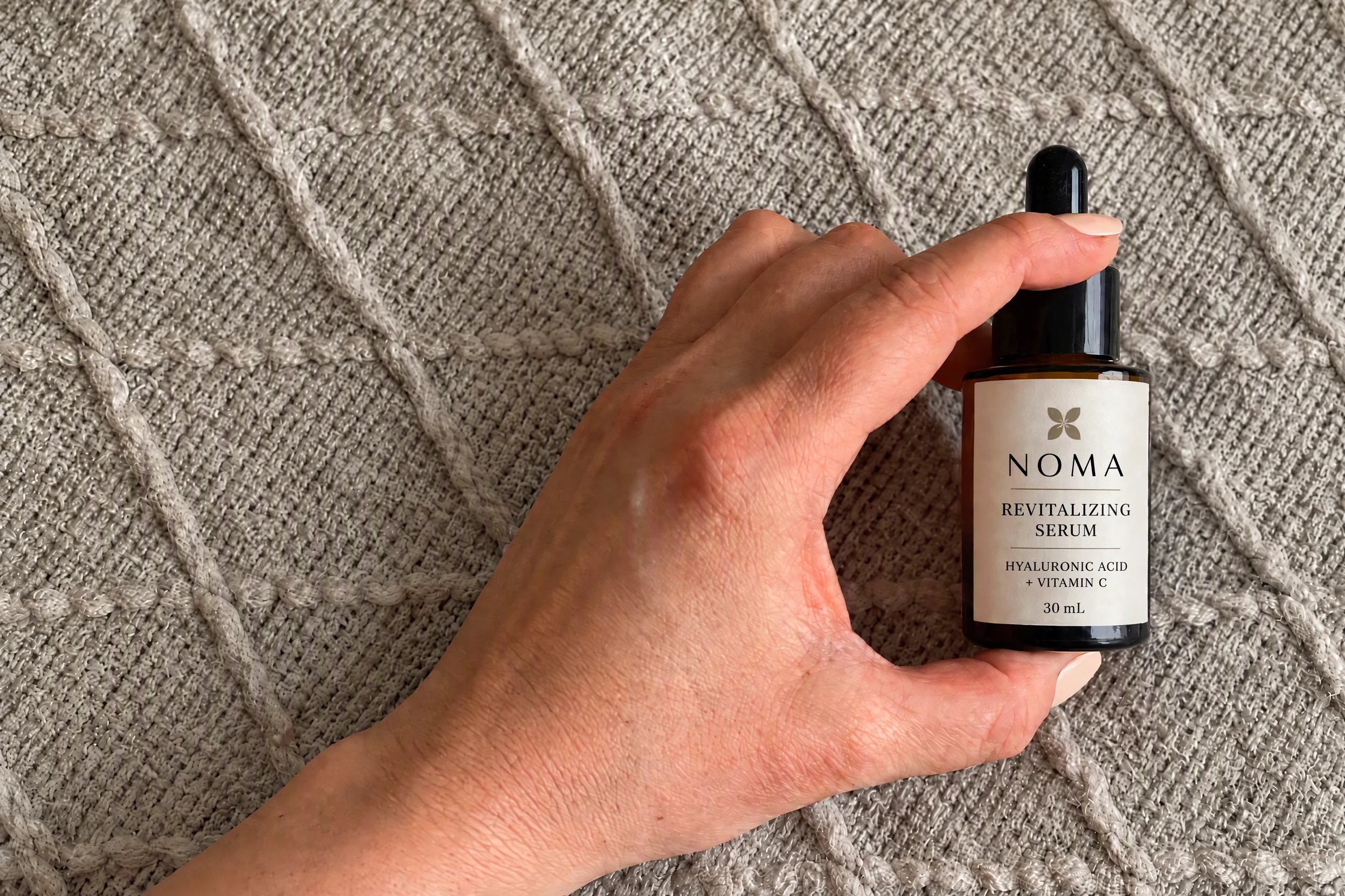 NOMA serum held above a knit blanket, based on a real phone photo with its demo label and wider framing edited in HummingBytes