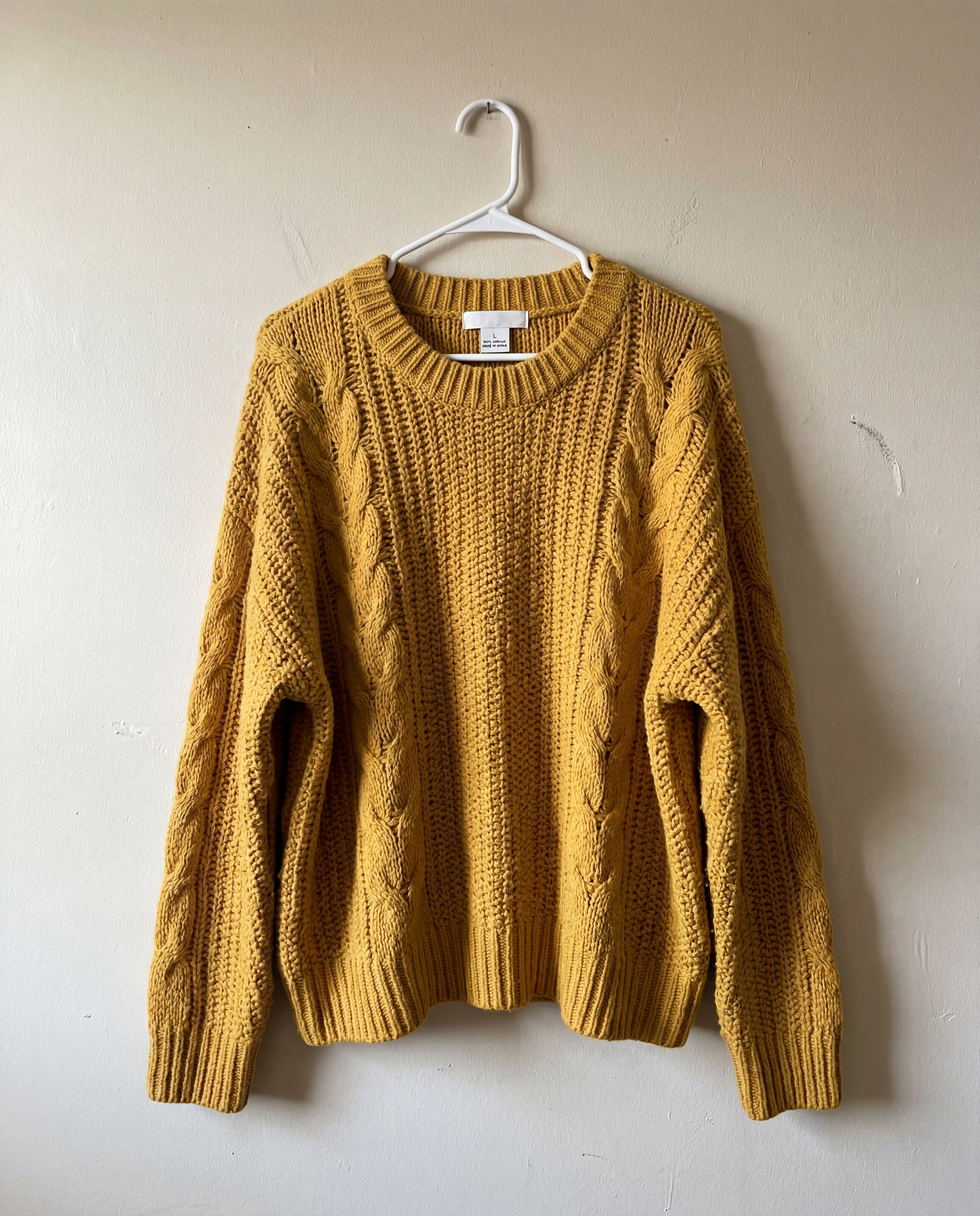 Raw smartphone-style source photo of a chunky mustard-yellow oversized sweater hanging on a plastic hanger against a beige wall